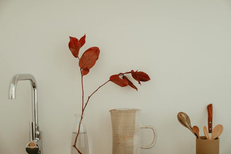 Elegant minimalist kitchen setup featuring a ceramic jug, red leaves, and cutlery on a white background.